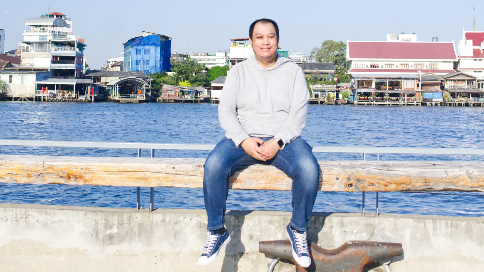 The man is sitting on the wall beside the Chao Phraya River under a clear sky.